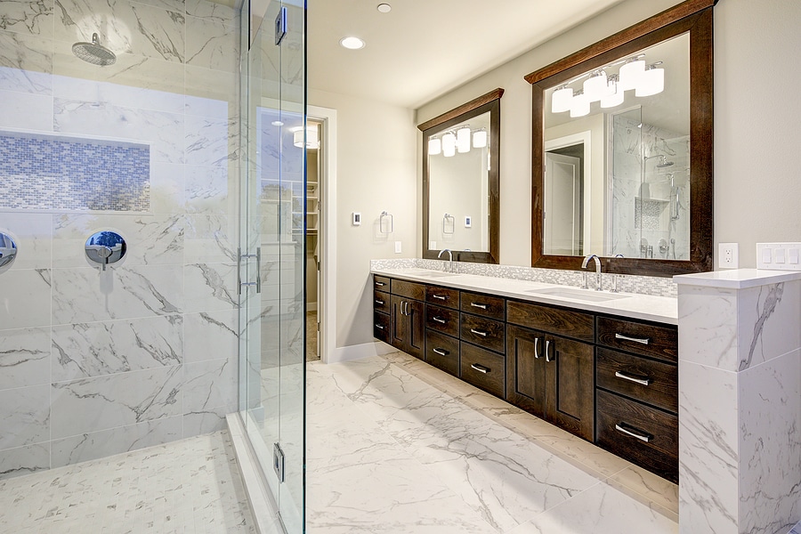 Photo of bright, airy master bathroom that features white walls, a modern, double vanity with rich brown cabinets and marble top, chrome fixtures throughout, and a marble walk-in shower with glass doors. 