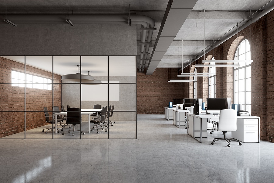 Open space office interior with brick and glass walls a concrete floor and arch windows. A row of computer desks and a conference room.